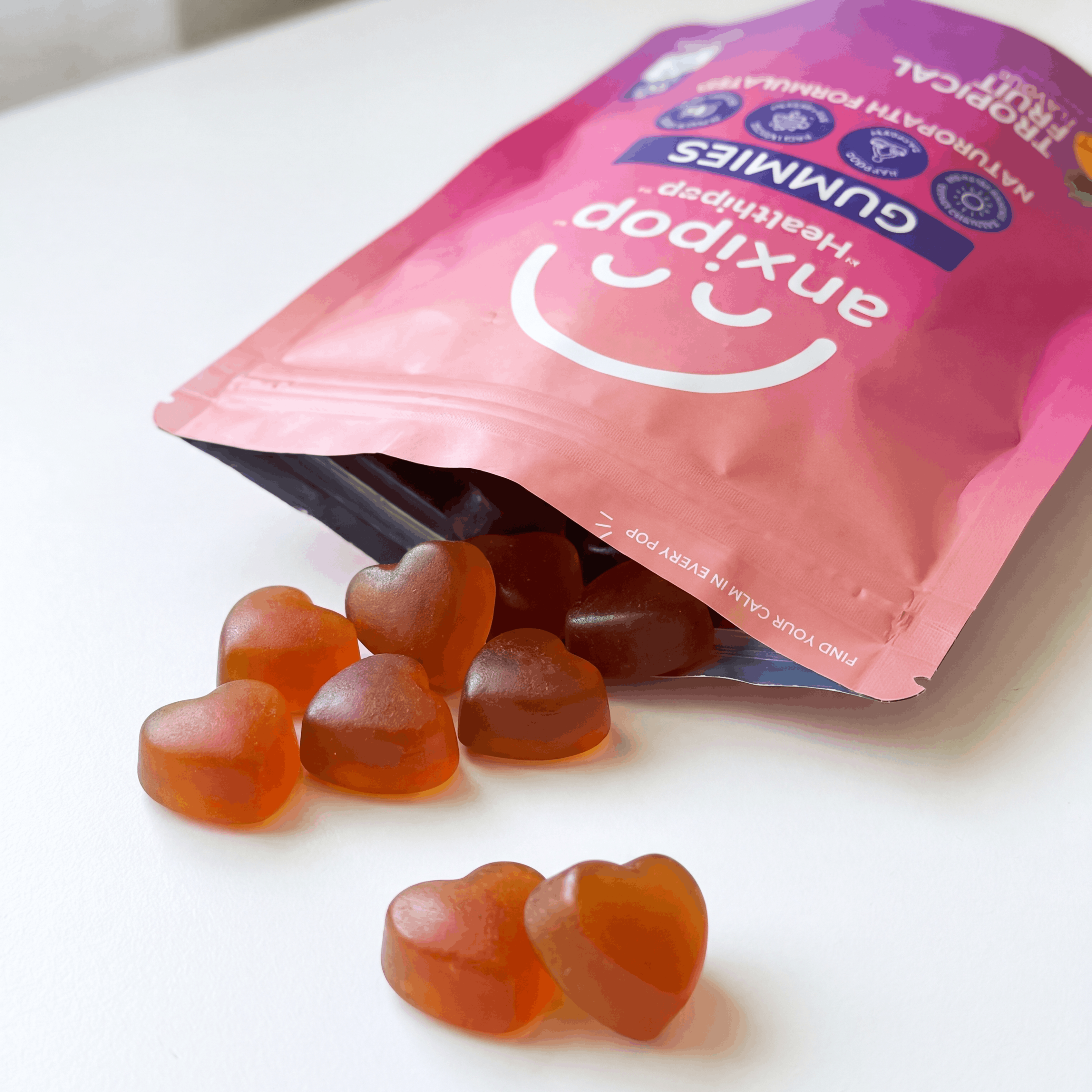 Anxipop heart-shaped saffron gummies, spilling out of a pink Anxipop resealable pouch on a white surface.